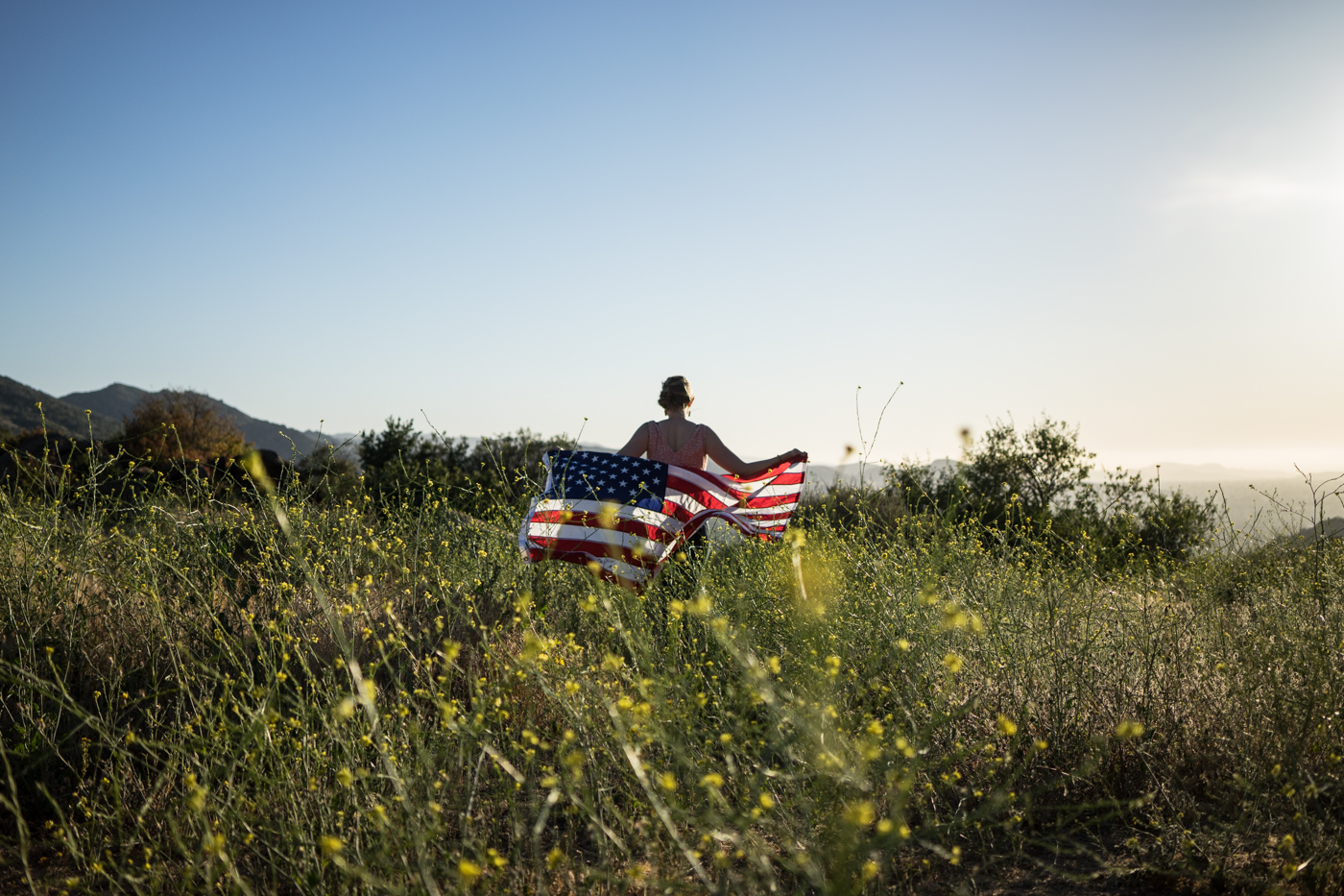 American woman with waving flag portrait in grassy wildflower meadow landscape at sunset
