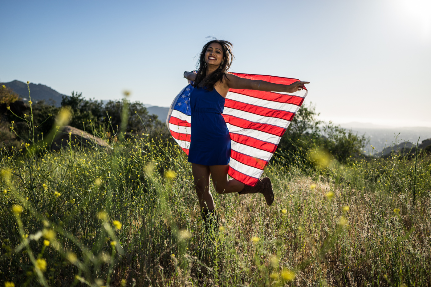 American woman jumping with flag portrait in grassy meadow landscape at sunset