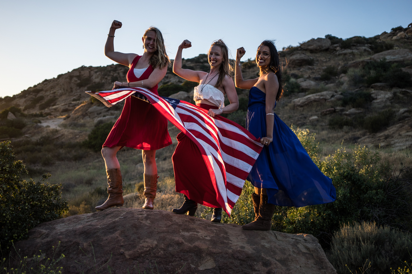 American women with waving flag portrait in grassy mountain landscape at sunset in rosy the riveter pose